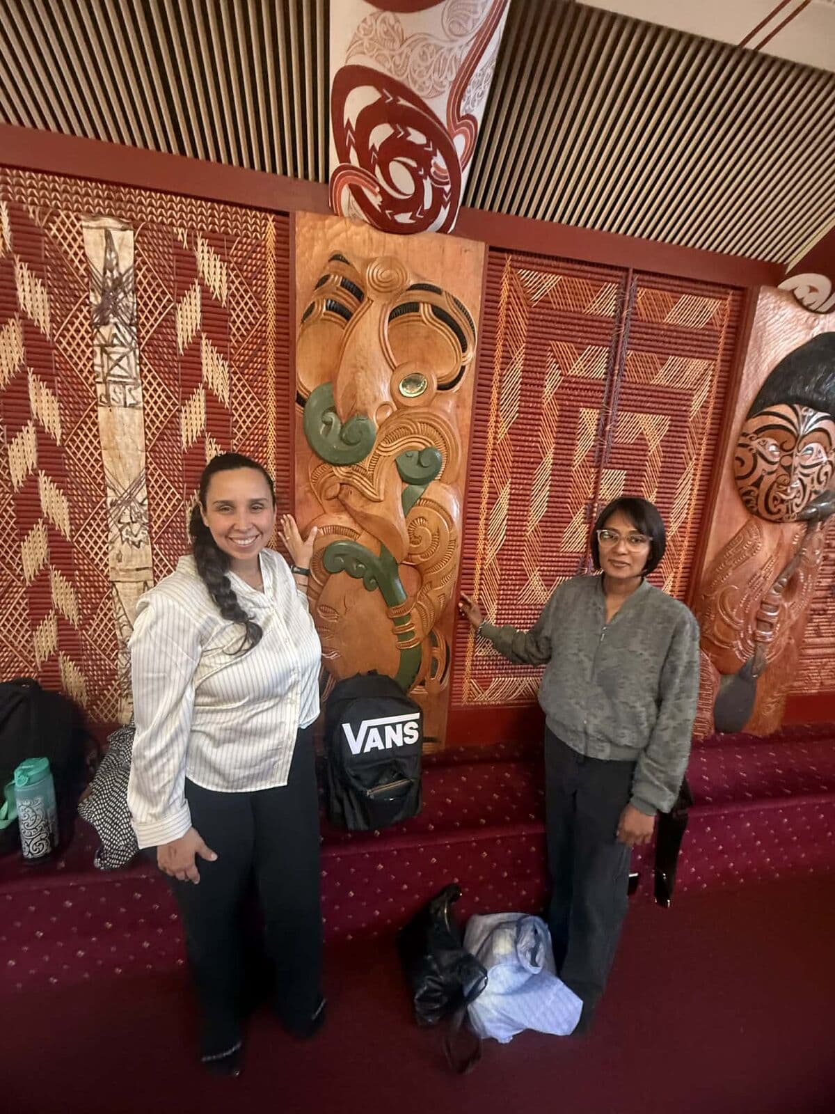 Two women standing beside whakairo with pounamu